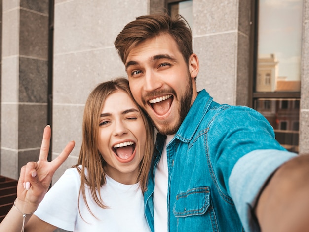 Free photo smiling beautiful girl and her handsome boyfriend in casual summer clothes. happy family taking selfie self portrait of themselves on smartphone camera. shows peace sign and winking in the street