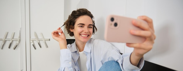 Бесплатное фото Улыбающаяся девушка делает селфи на смартфоне, позируя для фотографии в приложении для мобильного телефона, сидящем в ней