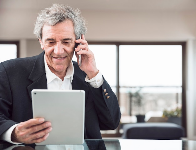 Free photo smiling senior man talking on mobile phone looking at digital tablet at workplace