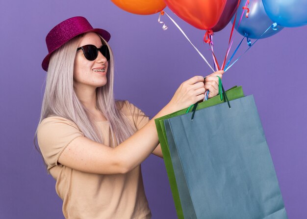 Free photo smiling young beautiful girl wearing dental braces and party hat with glasses holding balloons with gift bag isolated on blue wall
