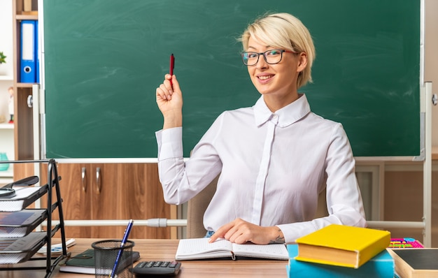 Бесплатное фото Улыбающаяся молодая блондинка учительница в очках, сидя за столом со школьными инструментами в классе, указывая пальцем на блокнот, глядя на фронт, указывая на классную доску с ручкой