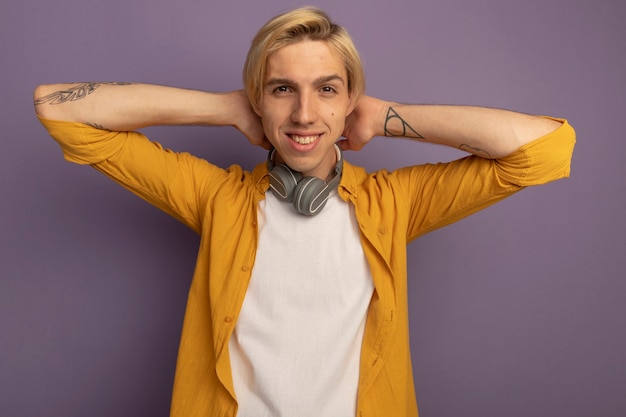 Бесплатное фото Улыбающийся молодой блондин в желтой футболке и наушниках на шее, держащий руки на шее, изолированный на фиолетовом