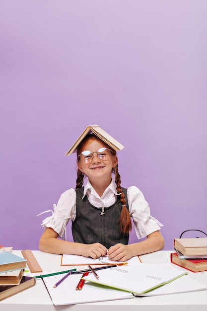 Бесплатное фото Улыбающаяся юная школьница с рыжими косичками в очках и книгой на голове в классическом наряде позирует, сидя за партой