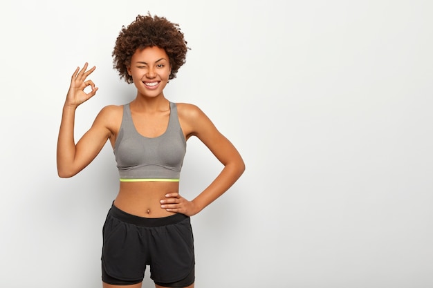 Бесплатное фото Спортивная модель в спортивной одежде показывает нормальный жест рукой, уверяет, что все в порядке, другую руку держит на талии