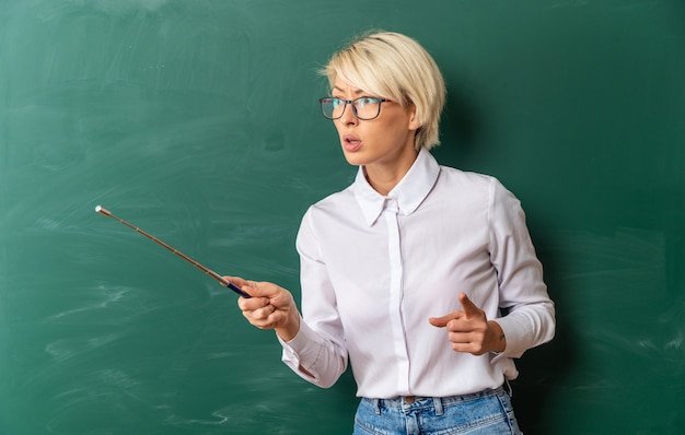 Бесплатное фото Строгая молодая блондинка учительница в очках в классе, стоя перед классной доской, глядя и указывая в сторону с указательной палкой и пальцем