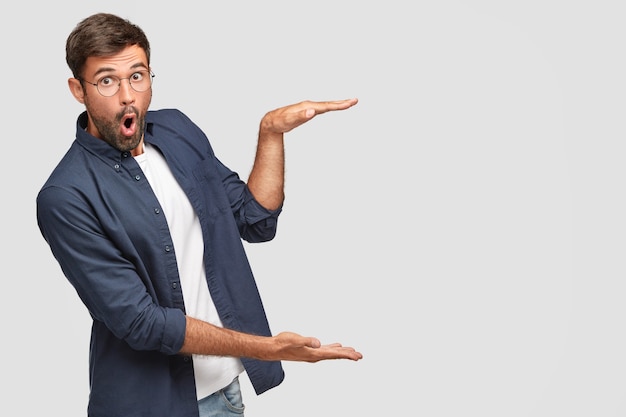 Free photo stunned unshaven male with shocked facial expression gestures with hands, shows size or height of something, dressed in fashionable shirt, isolated over white wall, copyspace