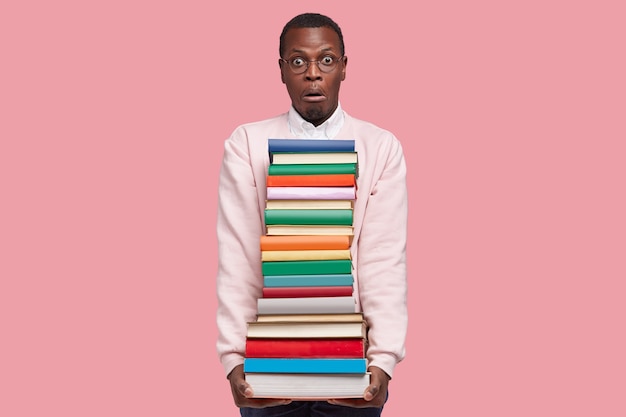 Ошеломленный молодой афроамериканец с большой стопкой книг, одетый в повседневный свитер, удивил выражение лица