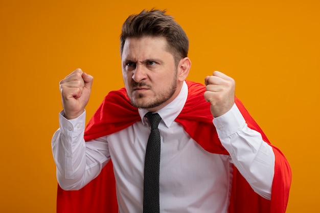 Бесплатное фото Супергерой бизнесмен в красном плаще сжимает кулак с сердитым лицом, стоящим над оранжевой стеной