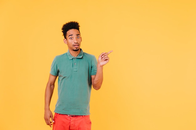 Free photo surprised man in stylish green tshirt pointing finger at something indoor shot of emotional brunette guy in casual outfit posing on yellow background
