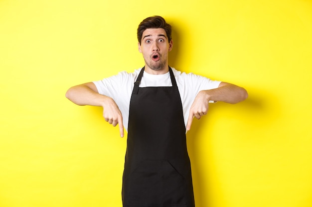 Free photo surprised seller in black apron pointing fingers down, looking amazed at camera, standing over yellow background.