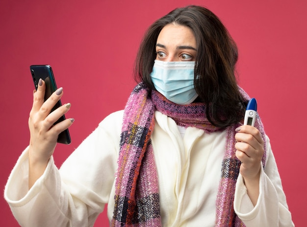 Бесплатное фото Удивленная молодая кавказская больная девушка в халате и шарфе с маской, держащая мобильный телефон и термометр, глядя на телефон, изолированный на малиновой стене