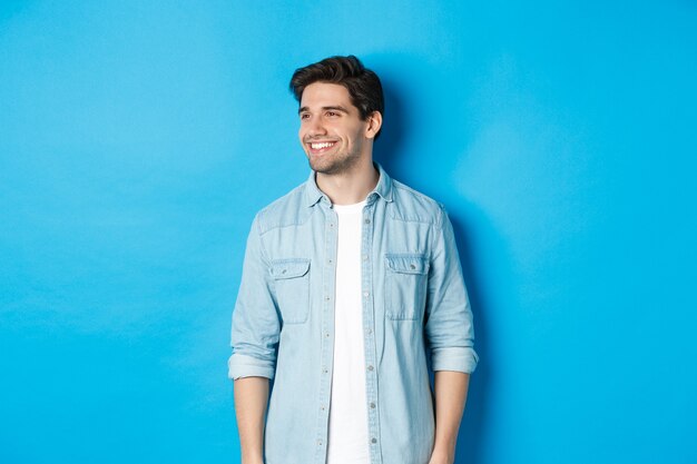 Bel homme adulte souriant en tenue décontractée, souriant et regardant à gauche l'offre promotionnelle, debout sur fond bleu.