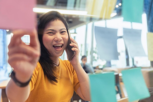 Photo gratuite une femme créative asiatique indépendante attrayante porte une conversation décontractée en tissu jaune avec un rappel de note de papier de point de main de smartphone dans un espace de coworking nouveau style de vie avec réflexion et fraîcheur