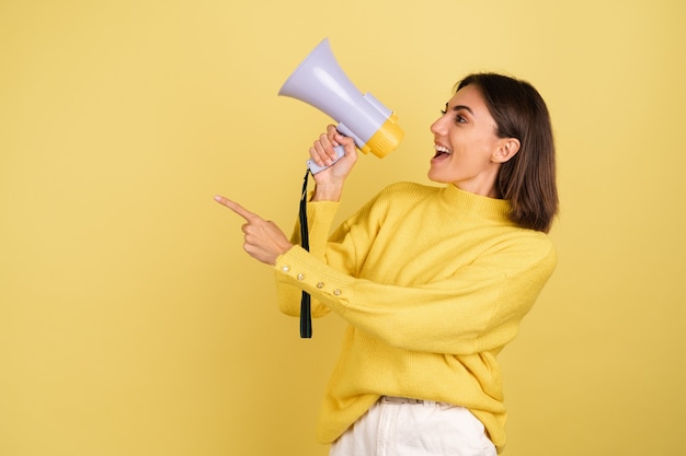 Photo gratuite jeune femme en pull chaud jaune avec haut-parleur mégaphone criant vers la gauche pointant l'index