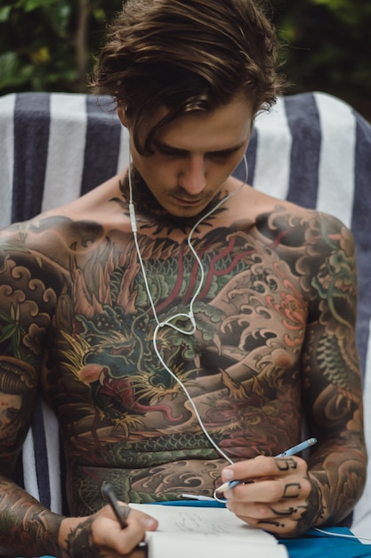 Photo gratuite un jeune homme tatoué portant des écouteurs écoute de la musique et dessine dans un cahier.
