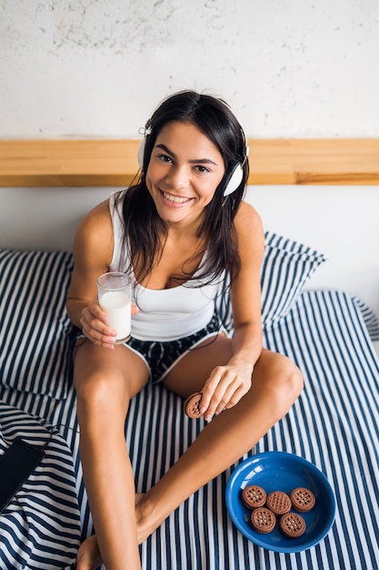 Photo gratuite jolie femme souriante sexy assise dans son lit le matin, écouter de la musique sur les écouteurs, prendre le petit déjeuner, manger des biscuits et boire du lait, mode de vie sain