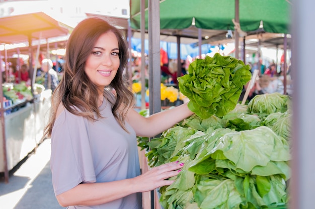 Photo gratuite portrait d'une belle jeune femme qui choisit des légumes à feuilles vertes dans un marché vert. concept de shopping alimentaire sain. jeune femme achetant des légumes au marché vert.