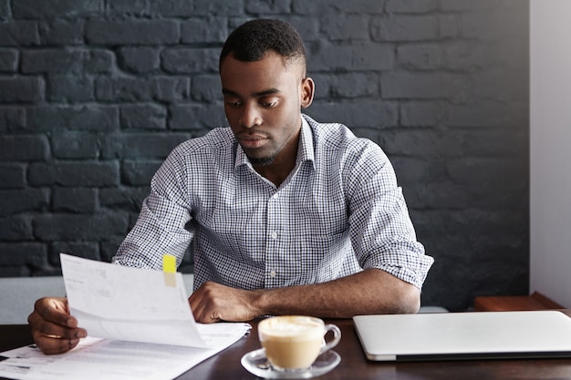 Photo gratuite portrait d'homme d'affaires afro-américain en chemise à carreaux examinant de près un morceau de papier dans ses mains