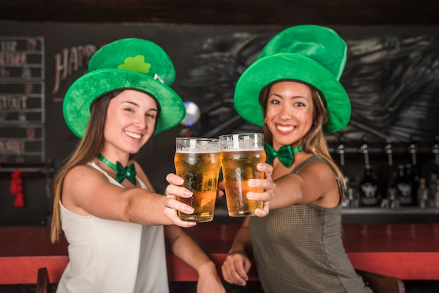 Rire de jeunes femmes en chapeaux Saint Patrick montrant des verres de boisson au comptoir