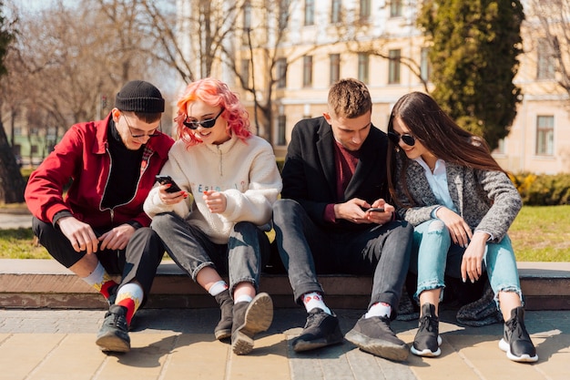 Vue de face de quatre amis ensemble à l'extérieur vérifiant leurs smartphones