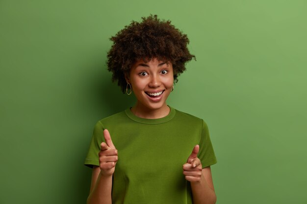 Photo gratuite vérifiez-le. sympathique jeune femme afro-américaine positive pointe l'index vers vous, choisit la personne et invite à rejoindre l'équipe, félicite ou loue le bon travail, isolé sur un mur vert