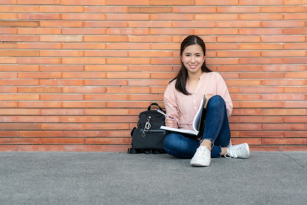 Photo Étudiant femme souriante tenant livre posant sur fond de brique sur le campus.