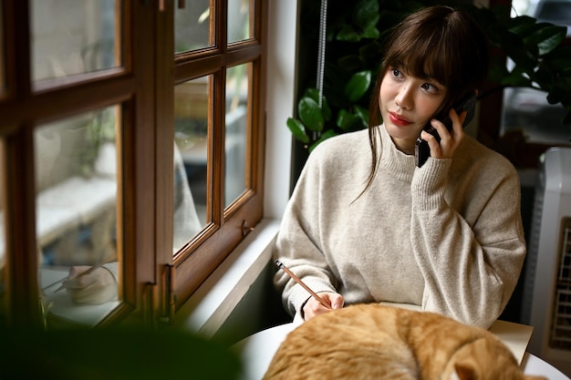 Une femme en pull douillet parle au téléphone assise à une table près de la fenêtre avec son chat