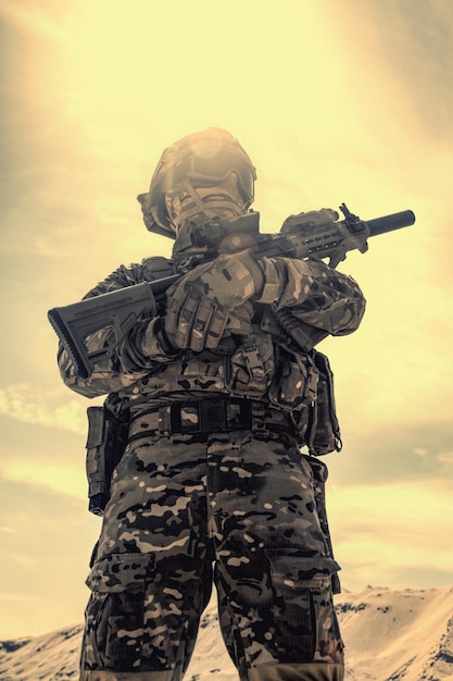 Joueur d'airsoft en uniforme de l'armée américaine, casque, masque et lunettes debout avec réplique de fusil de service dans une zone sablonneuse à faible angle, tir désaturé. Jeux de guerre, participant à la reconstitution militaire dans le désert