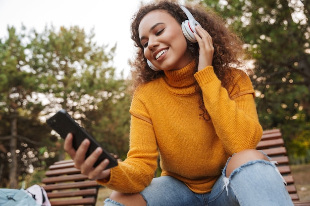 Photo photo d'une belle jeune femme bouclée optimiste joyeuse s'asseoir sur un banc dans un parc à l'extérieur en écoutant de la musique avec des écouteurs à l'aide d'un téléphone portable.