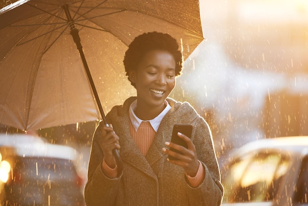 Photo d'une jeune femme d'affaires utilisant un parapluie et un smartphone tout en se promenant sous la pluie dans un contexte urbain