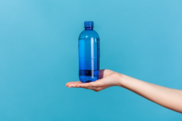 Bliska kobieca ręka demonstruje plastikową niebieską butelkę z wodą mineralną, reklama świeżego czystego napoju, najlepszy sposób na przywrócenie równowagi elektrolitów. Kryty studio strzał na białym tle na niebieskim tle