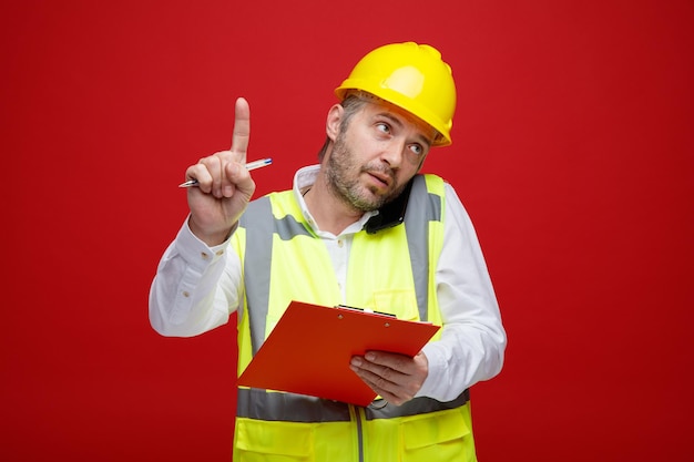 Konstruktor w mundurze budowlanym i kasku ochronnym, trzymając schowek, rozmawiając na telefonie komórkowym, wyglądający pewnie, prosząc o czekanie pokazując palec wskazujący stojący na czerwonym tle