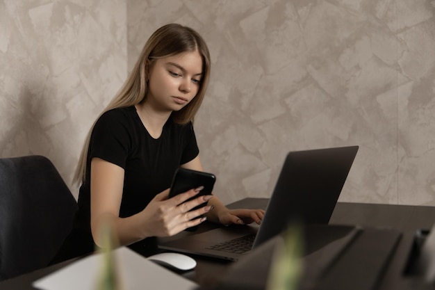 Piękna dziewczyna pracuje zdalnie w domu przy komputerze i sprawdza powiadomienia przez telefon