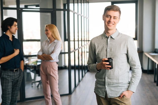 Zdjęcie portret pozytywnego młodego pracownika biurowego w przypadkowych ubraniach trzymającego filiżankę kawy na wynos w ręku stojącego w biurze uśmiechającego się patrzącego w kamerę