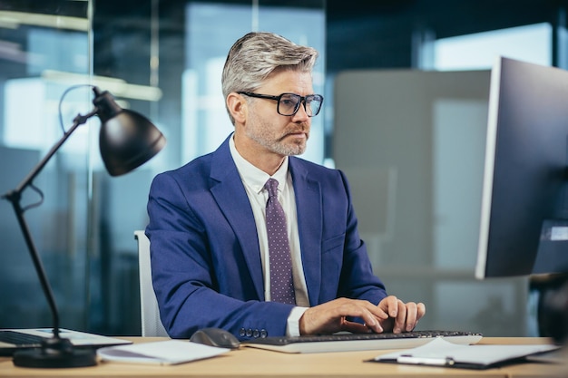 Zdjęcie poważny menedżer w okularach siwy z brodą pracujący przy komputerze w nowoczesnym biurze biznesowy mężczyzna w pracy