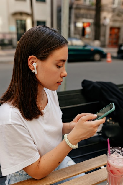 Stylowa piękna dziewczyna z rozpuszczonymi ciemnymi włosami siedząca na zewnątrz ze smartfonem w bezprzewodowych słuchawkach i przewijanym smartfonem