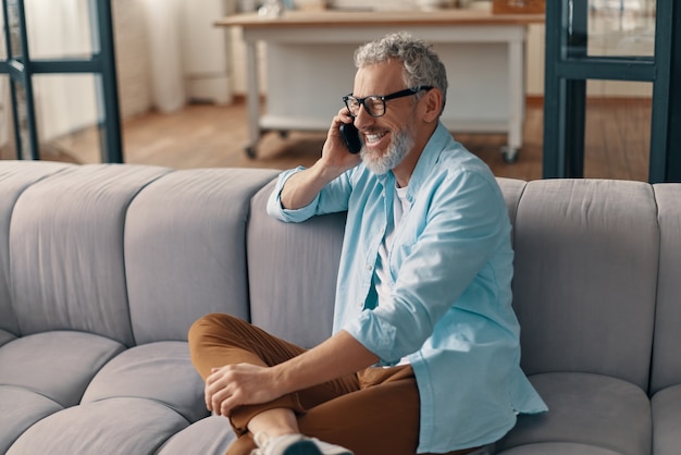 Szczęśliwy starszy mężczyzna w swobodnej odzieży i okularach rozmawia przez smartfon siedząc na kanapie w domu
