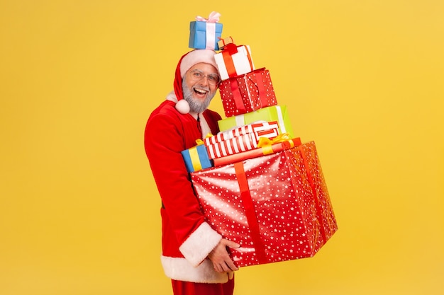 Szczęśliwy zabawny starszy mężczyzna z siwą brodą w stroju Świętego Mikołaja trzymając stos świątecznych prezentów w rękach i na głowie, świętując nowy rok. Kryty studio strzał na białym tle na żółtym tle.