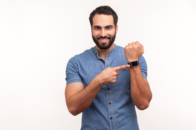 Wesoły pozytywny mężczyzna z brodą w niebieskiej koszuli, wskazujący na zegarek na rękę i uśmiechnięty, pokazujący nowy inteligentny zegar, sprawdzający wskaźniki. Kryty studio strzał na białym tle