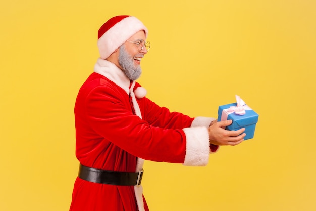 Widok z boku starszego mężczyzny z szarą brodą w stroju świętego mikołaja trzymającego pudełko prezentowe, powitanie w wigilię Bożego Narodzenia i nowego roku. Kryty studio strzał na białym tle na żółtym tle.
