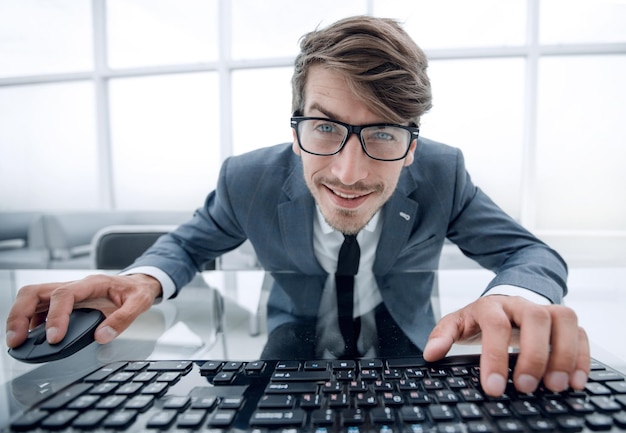 Zdjęcie zabawny młody człowiek z szaloną twarzą siedzący przy stole z rękami na myszy i klawiaturze komputera