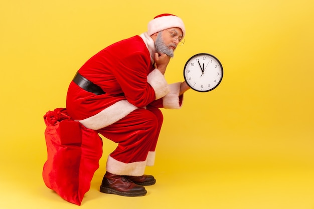 Znudzony starszy mężczyzna ubrany w strój Świętego Mikołaja siedzący na dużej czerwonej torbie z prezentami i trzymający w dłoni zegar ścienny, czekający na prezent. Kryty studio strzał na białym tle na żółtym tle.