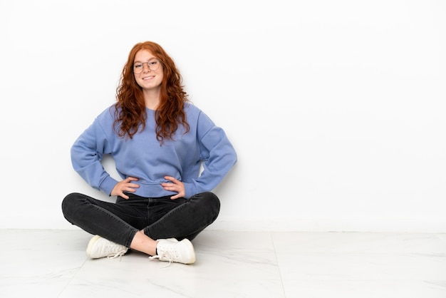 Photo teenager redhead girl sitting on the floor isolated on white background posing with arms at hip and smiling