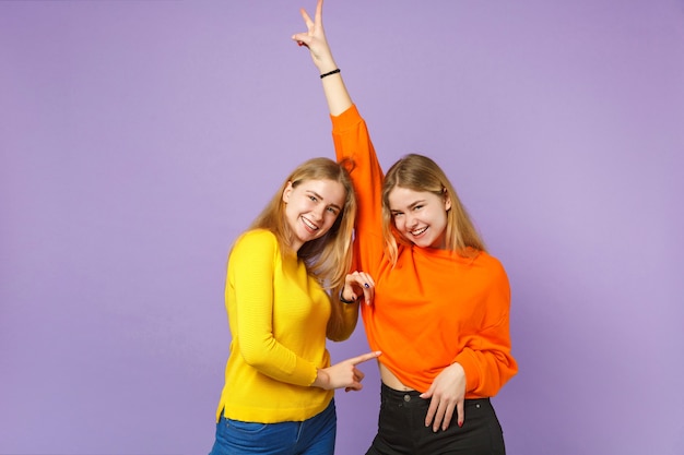 Фото Две смеющиеся молодые блондинки сестры-близнецы девушки в красочной одежде, показывая знак победы, изолированные на пастельной фиолетовой синей стене. концепция семейного образа жизни людей.