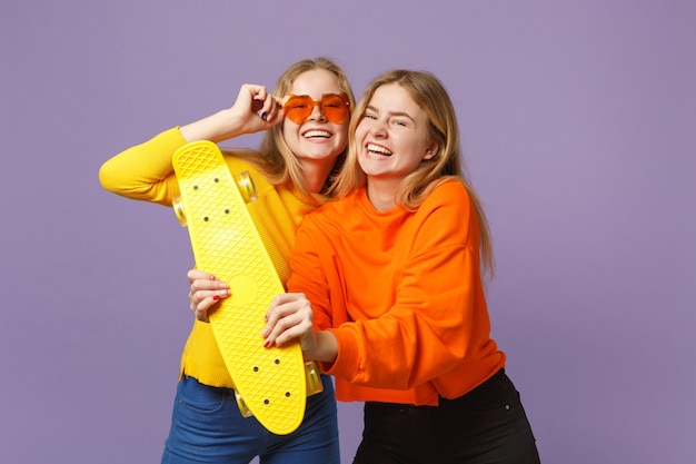 Фото Две смеющиеся молодые белокурые девушки сестры-близнецы в яркой одежде, сердечные очки держат желтый скейтборд, изолированный на пастельно-фиолетовой синей стене. концепция семейного образа жизни людей.
