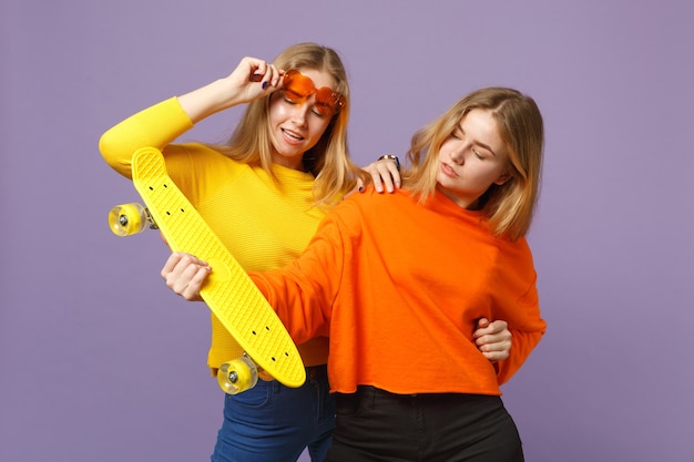 Фото Две потрясающие молодые блондинки сестры-близнецы в яркой одежде, в очках в форме сердца держат желтый скейтборд, изолированный на пастельно-фиолетовой синей стене. концепция семейного образа жизни людей. .