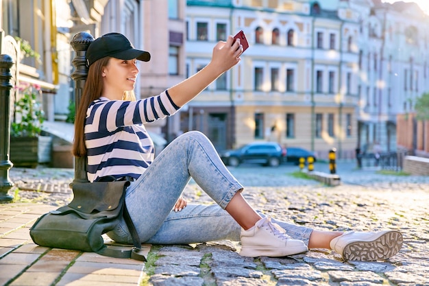 Модная девушка-подросток в городском стиле сидит на тротуаре с помощью смартфона