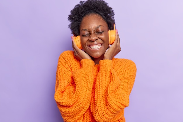  woman with curly hair keeps hands on headphones enjoys good sound pleasant melody dressed in orange knitted jumper isolated on purple 