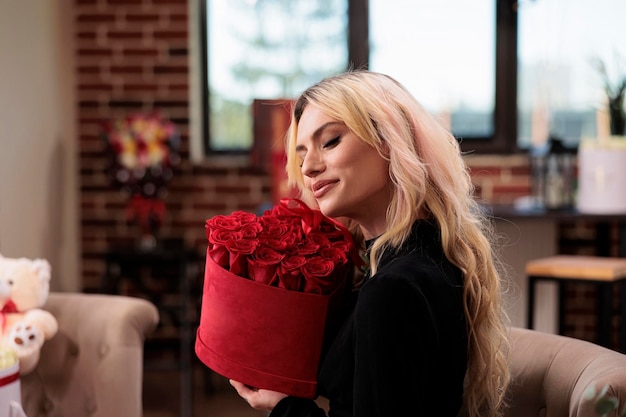 Woman holding valentines day red roses box, receiving luxury presents. Attractive blonde girlfriend with flower bouquet basket sitting in living room with expensive gifts, surprise.