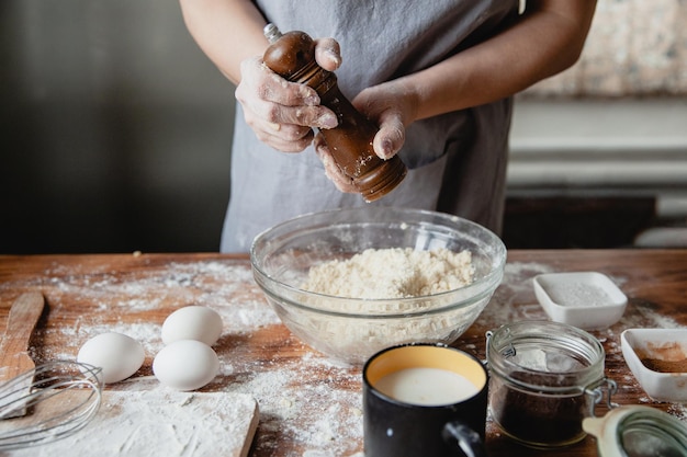 Женщина солит тесто в стеклянной миске на деревянном столе с помощью мельницы на кухне. На столе другие продукты.